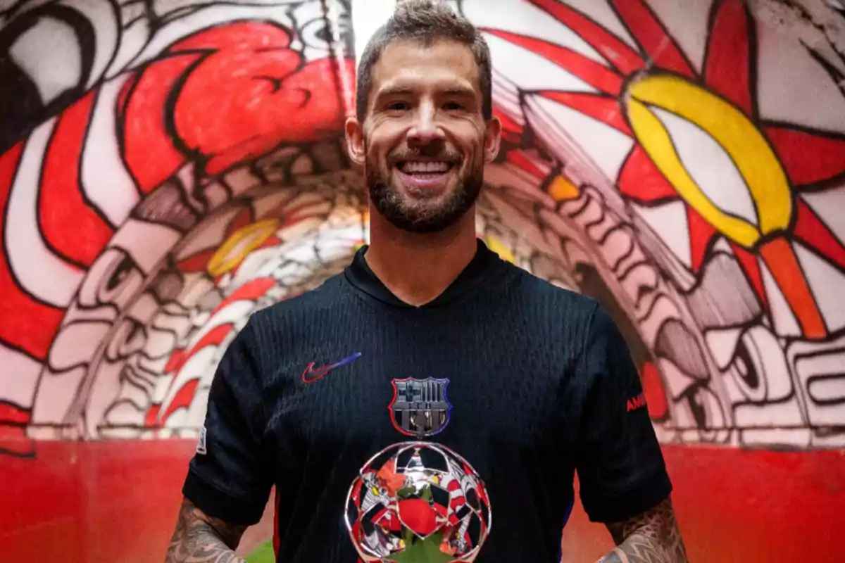 A smiling man in a black soccer jersey holds a trophy in front of a colorful mural. A smiling man in a black soccer jersey holds a trophy in front of a colorful mural.