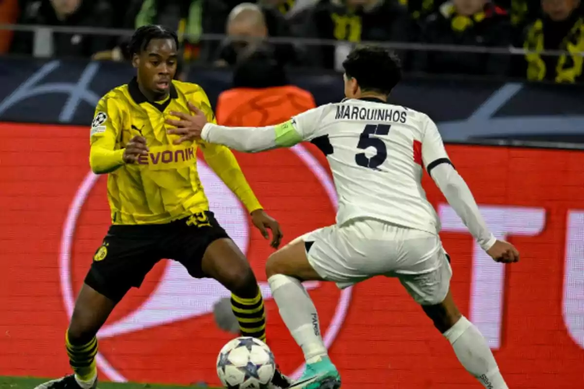 Dos jugadores de fútbol compiten por el balón durante un partido, uno con uniforme amarillo y negro y el otro con uniforme blanco.