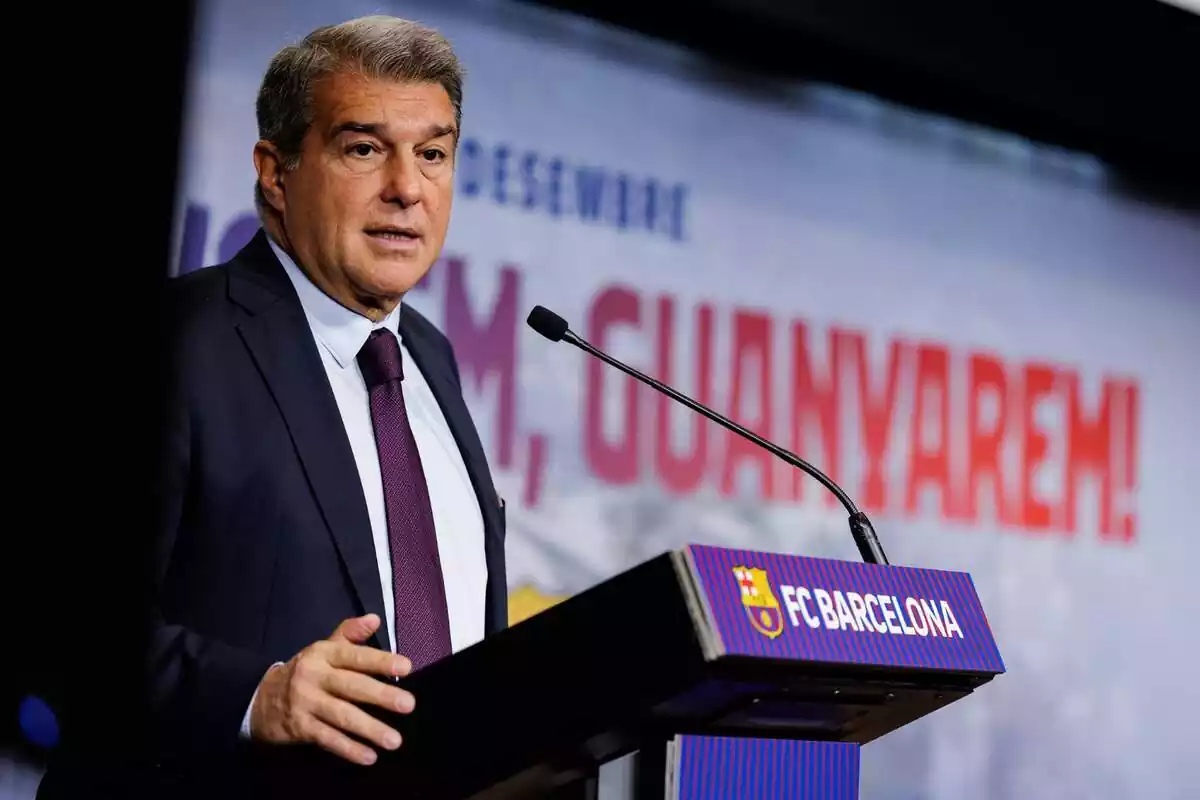 Joan Laporta, presidente del FC Barcelona, en una conferencia, vestido de terno y una corbata de colores azulgrana. En su delante, un micrófono moderno y un soporte de la institución deportiva FC Barcelona. Detrás, un fondo difuminado con un lema característico en catalán.