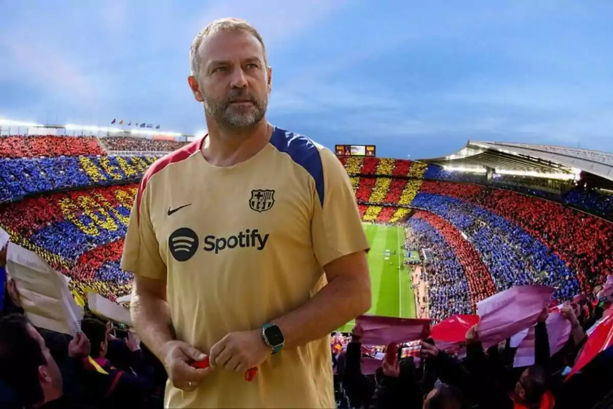 Un hombre con una camiseta de entrenamiento del FC Barcelona está de pie frente a un estadio lleno de aficionados que sostienen carteles de colores rojo, azul y amarillo.