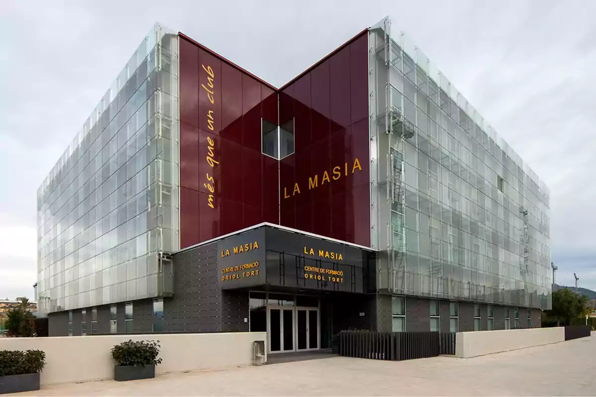 Edificio moderno de La Masia con fachada de vidrio y paneles rojos, con el lema "més que un club" en el lateral.