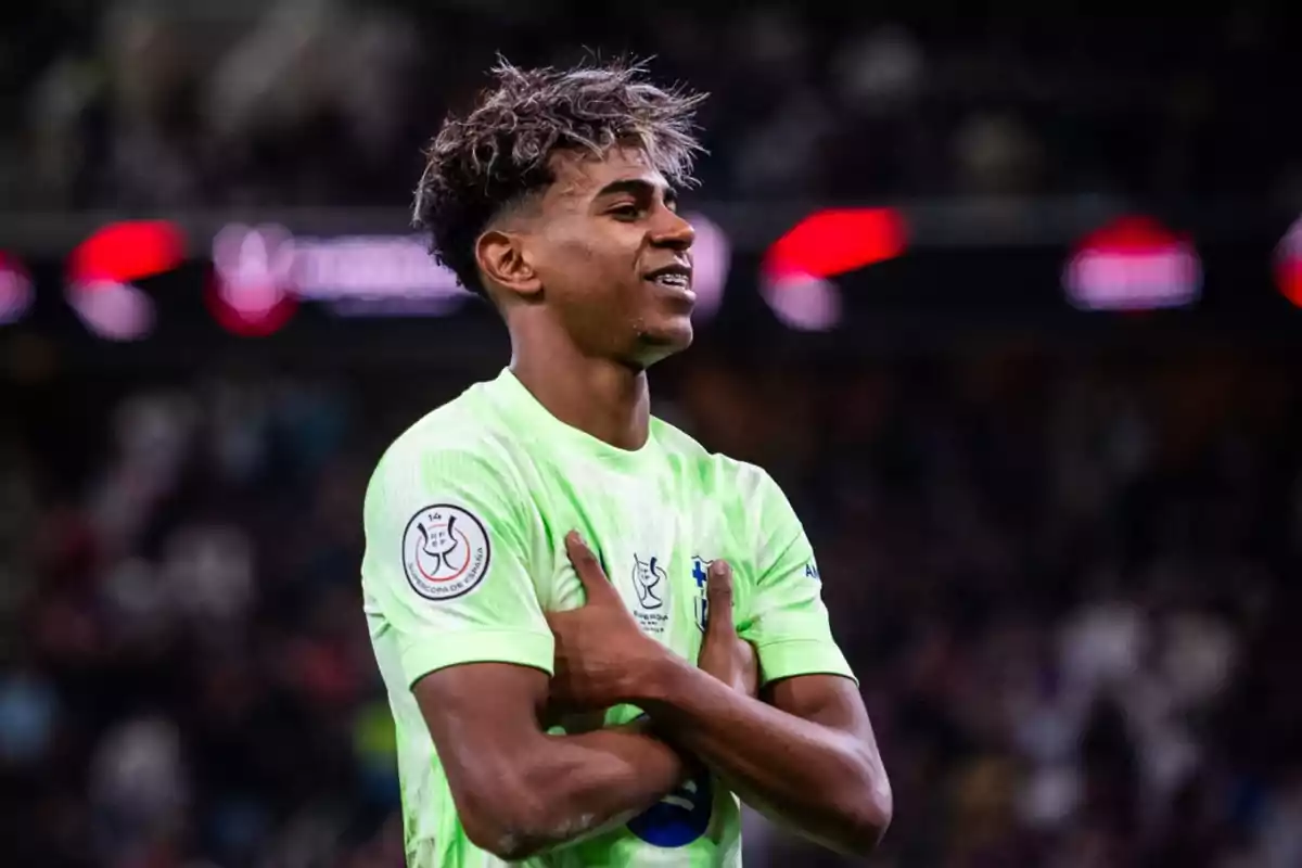 A soccer player in a green uniform celebrates on the field with his arms crossed over his chest.