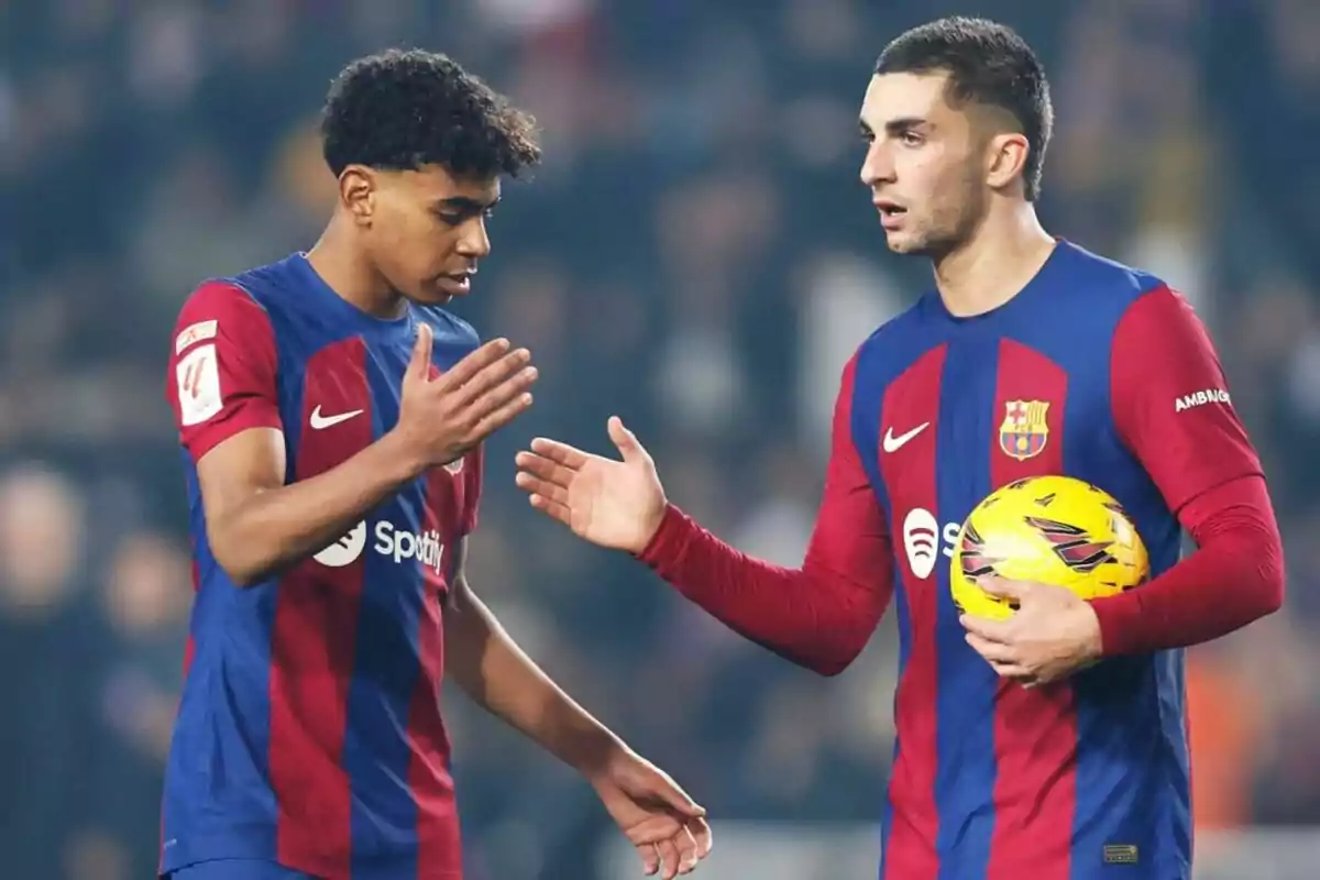 Dos jugadores de fútbol del FC Barcelona en el campo, uno sosteniendo un balón y el otro extendiendo la mano.