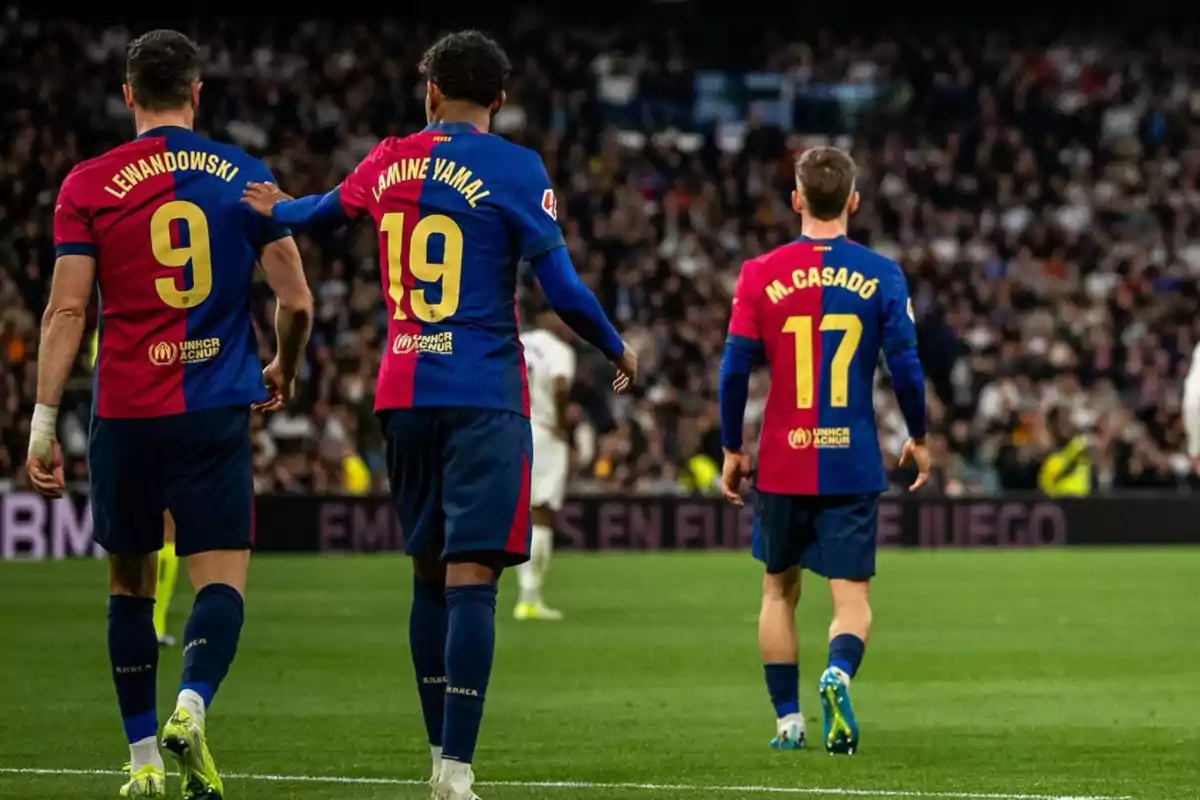 Tres jugadores de fútbol con uniformes azul y rojo caminan por el campo durante un partido.