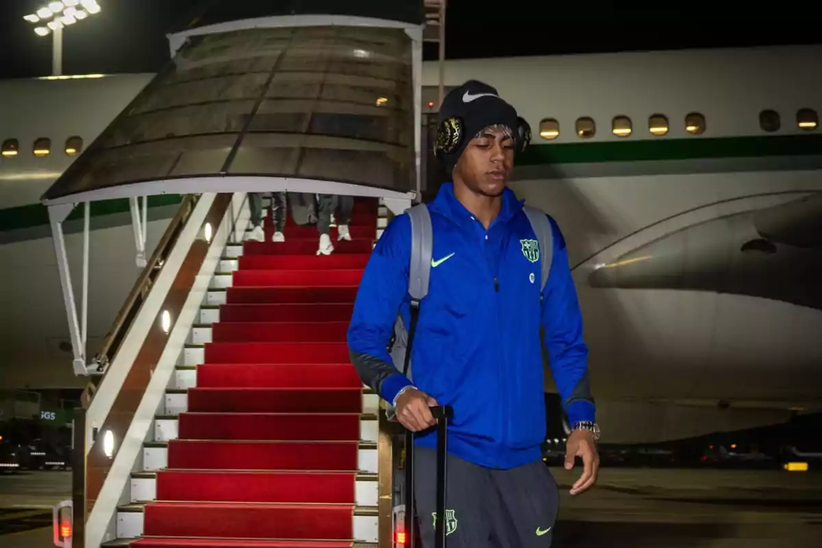 A man wearing headphones and blue sportswear steps off a plane down a red-carpeted staircase.