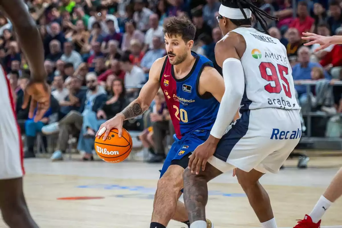 Un jugador de baloncesto con uniforme azul dribla el balón mientras es defendido por un jugador con uniforme blanco en una cancha llena de espectadores.