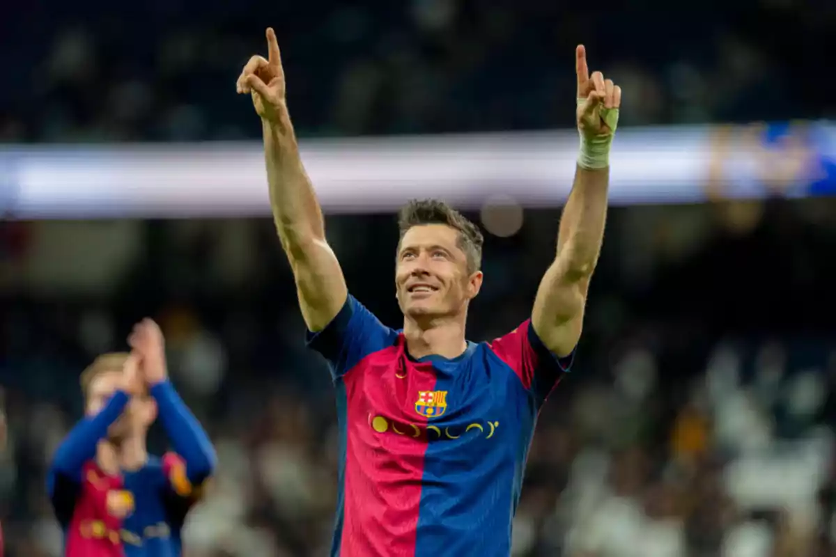 Un jugador de fútbol con uniforme azul y rojo levanta los brazos en señal de celebración en un estadio.