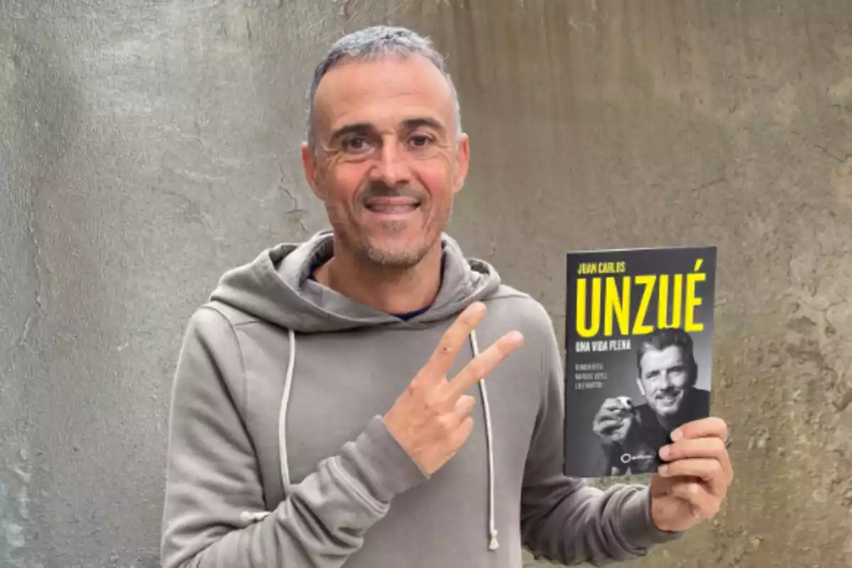 Aparece un hombre con una sudadera gris, con la mano derecha levantada con dos dedos y con la otra mano tiene un libro en el que se puede leer la palabra unzue