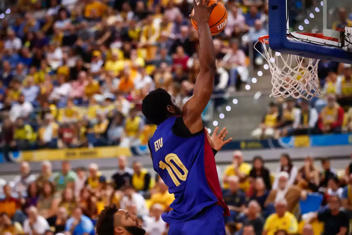 Jugador de baloncesto con el número 10 en la camiseta azul y roja realizando un mate durante un partido con público en las gradas.