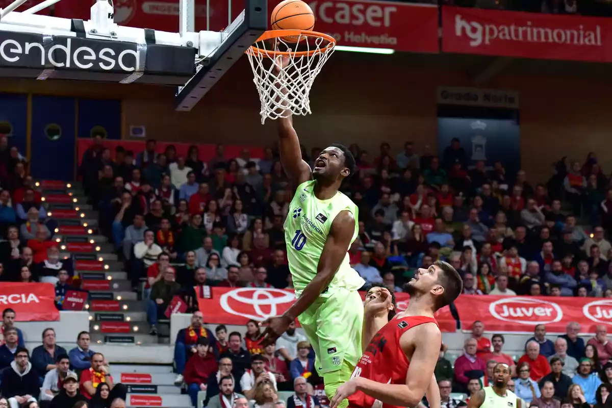 Un jugador de baloncesto con uniforme verde realiza una bandeja durante un partido, mientras un oponente con uniforme rojo intenta defenderlo, con una multitud de espectadores en el fondo.