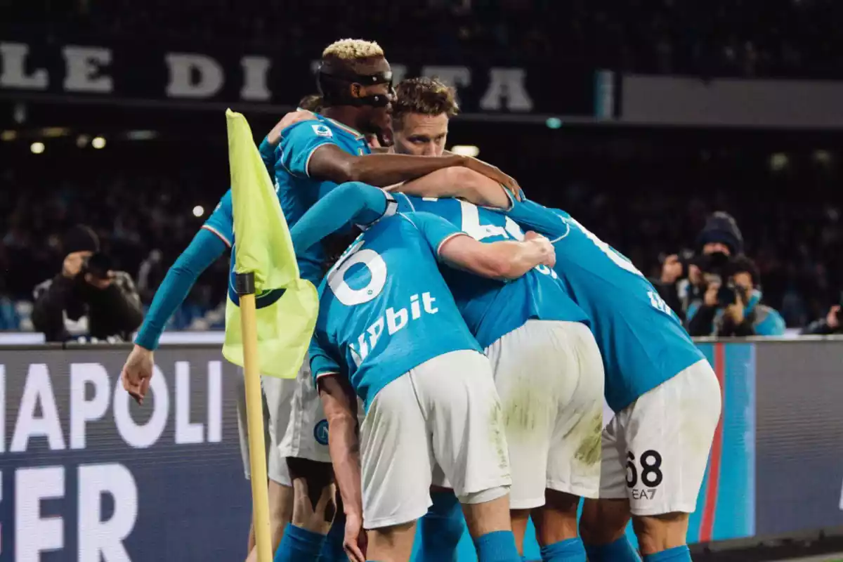 Jugadores del Napoli abrazados en el córner celebrando el gol con la camiseta celeste y los pantalones blancos