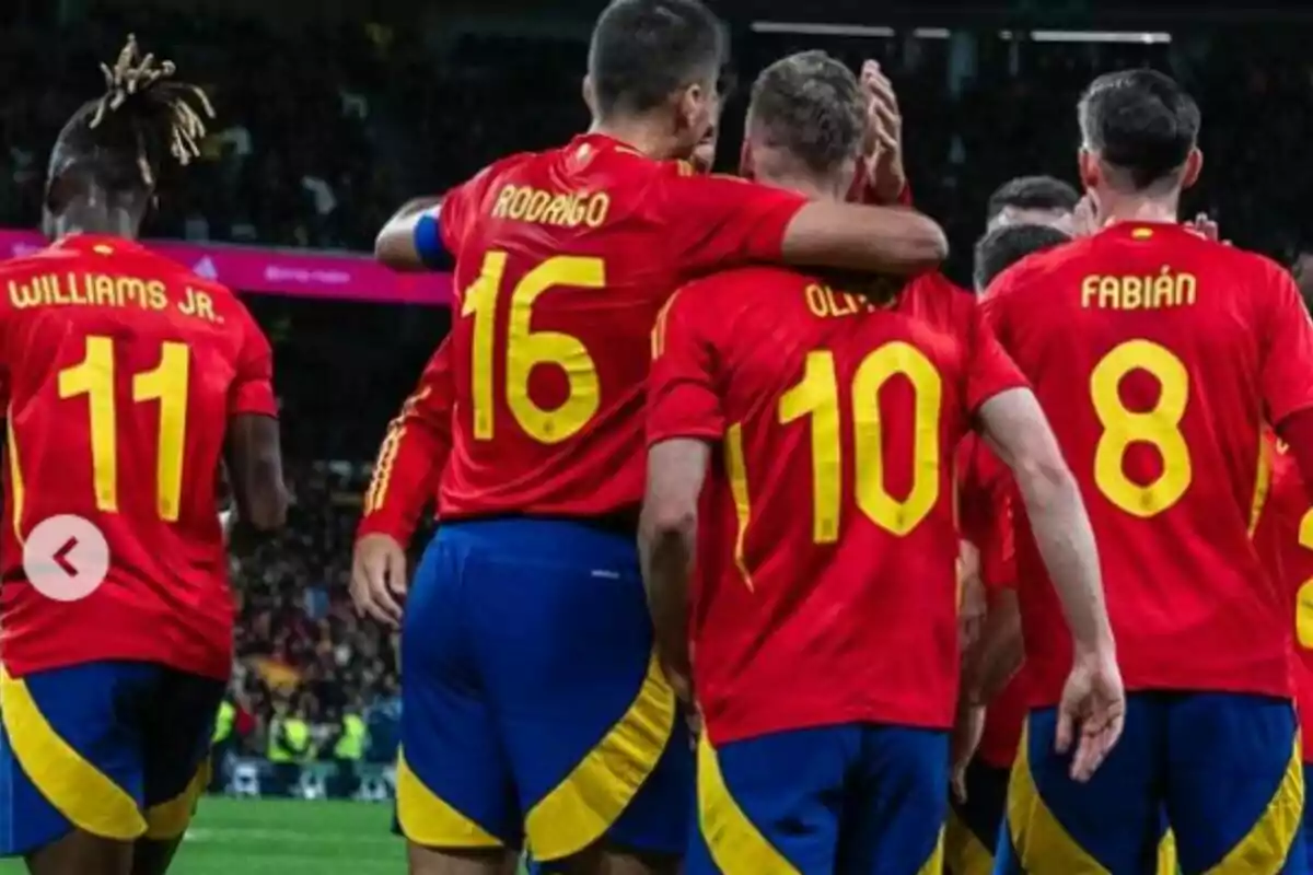 Jugadores de fútbol con uniformes rojos y azules celebran en el campo. Los nombres en sus camisetas son "Williams Jr.", "Rodrigo", "Olmo" y "Fabián", con los números 11, 16, 10 y 8 respectivamente. Están abrazados y parecen estar celebrando un gol o una victoria.