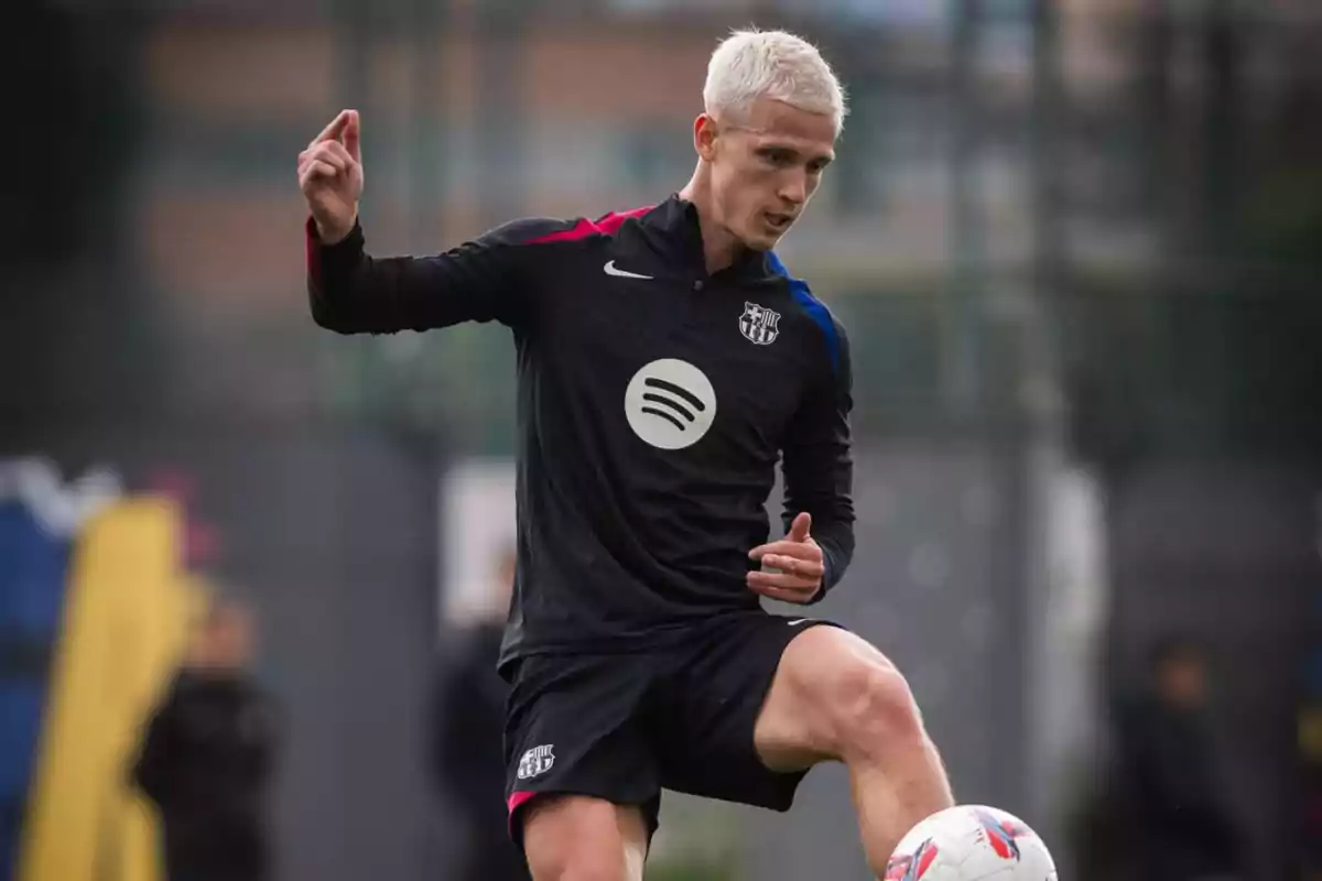 A soccer player with platinum blonde hair wears a black uniform with logos while controlling a ball on the field.