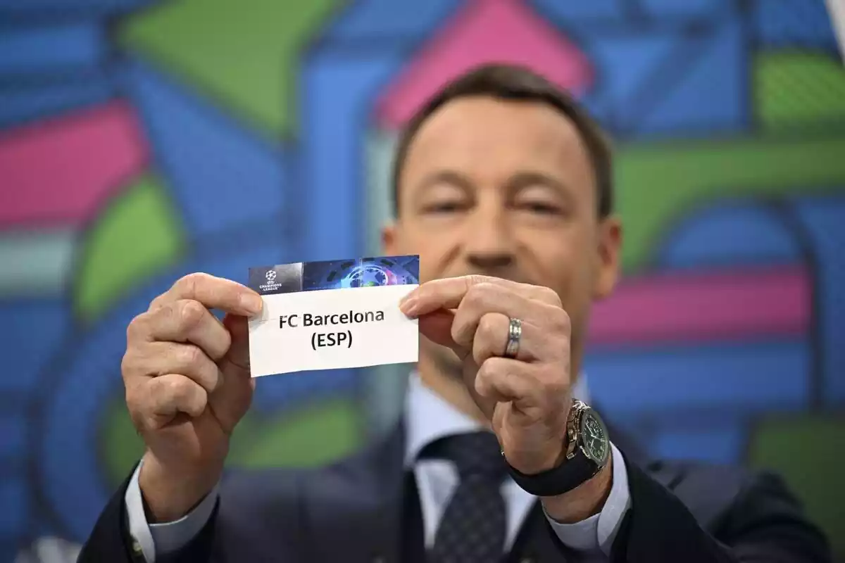Un hombre elegante sosteniendo un papel que dice "FC Barcelona (ESP)", en referencia al sorteo de la UEFA Champions League.