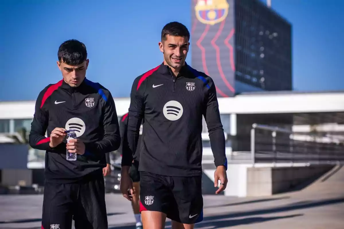 Two soccer players walking in black training clothes with a club and music brand logo in an urban setting.