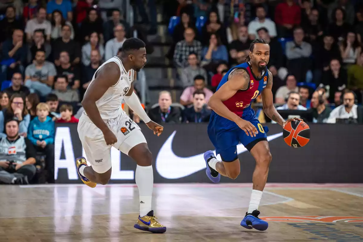 Dos jugadores de baloncesto en acción durante un partido, uno con uniforme azul y rojo dribla el balón mientras el otro con uniforme blanco lo persigue, con espectadores en el fondo.