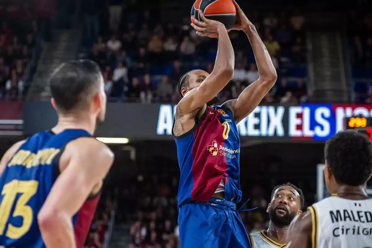 Un jugador de baloncesto del equipo con uniforme azul y rojo lanza el balón mientras otros jugadores observan en una cancha llena de espectadores.