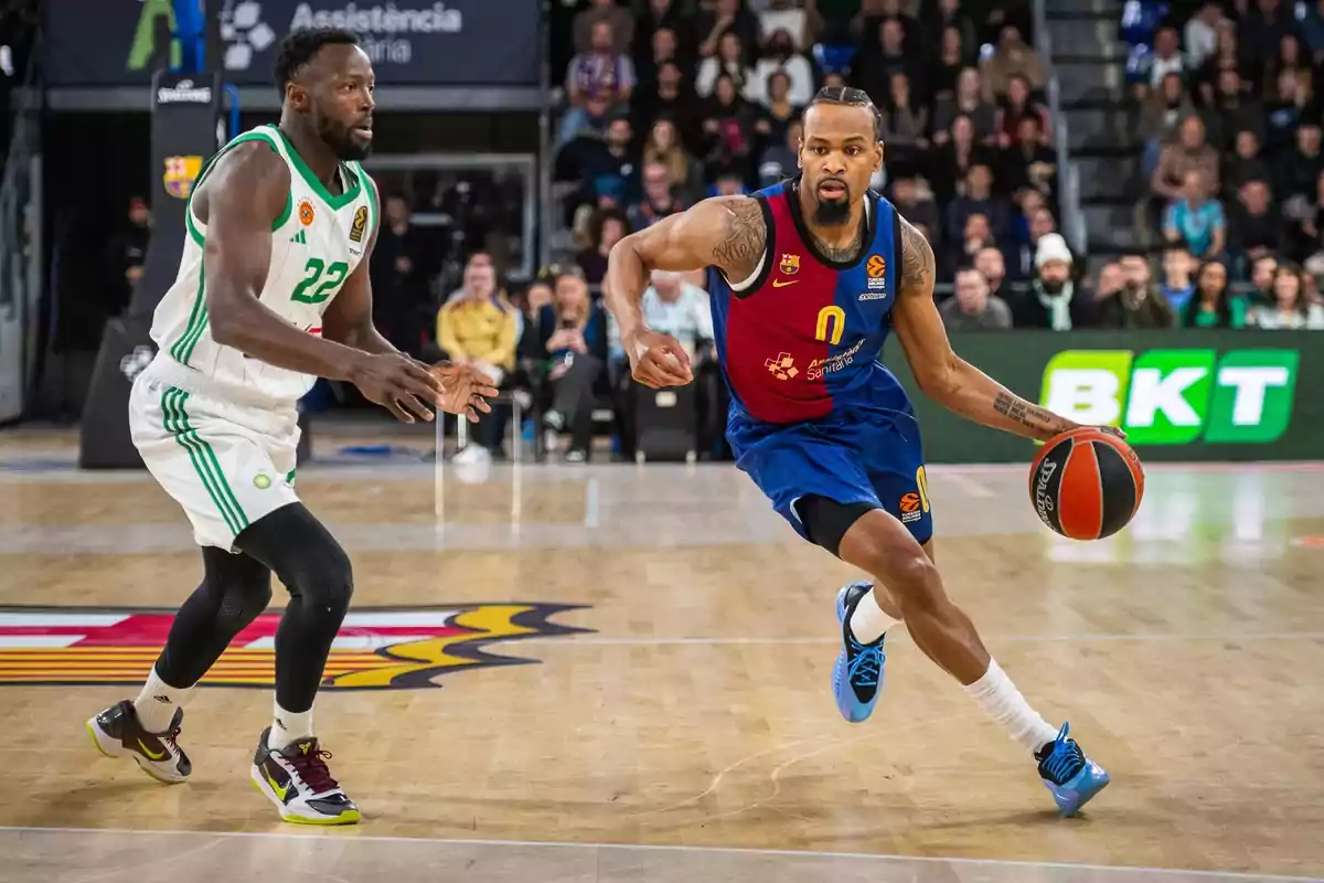 Dos jugadores de baloncesto en acción durante un partido en una cancha cubierta con público de fondo.