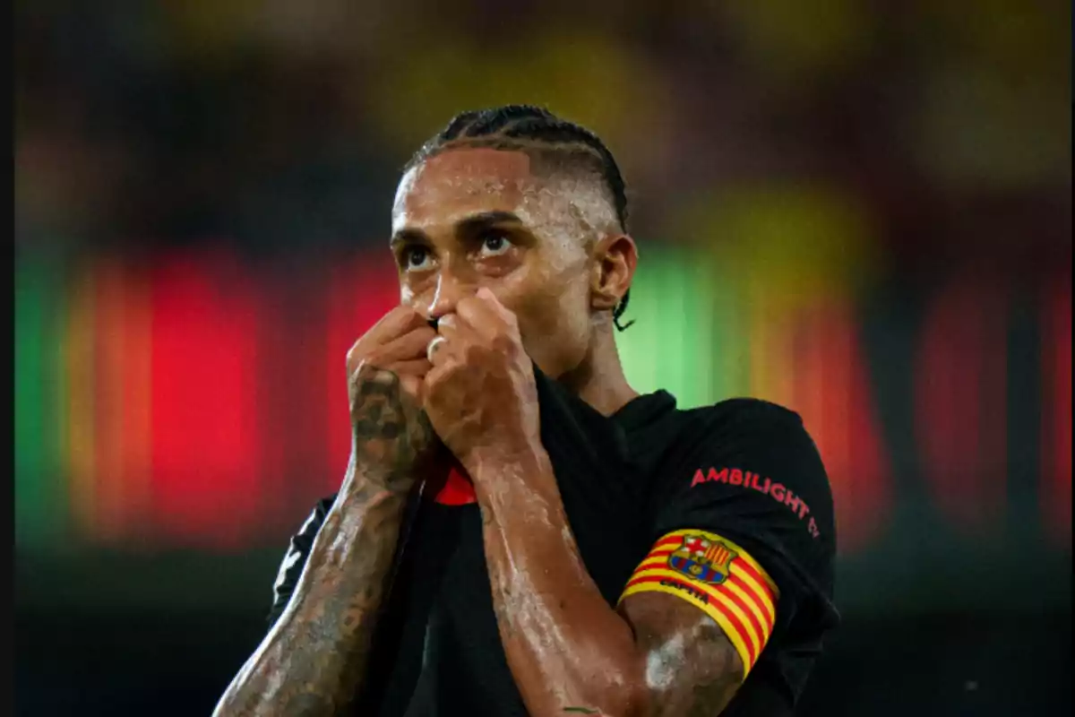 Un jugador de fútbol con una camiseta negra se cubre la boca con la camiseta mientras mira hacia arriba, lleva un brazalete de capitán con los colores del FC Barcelona.