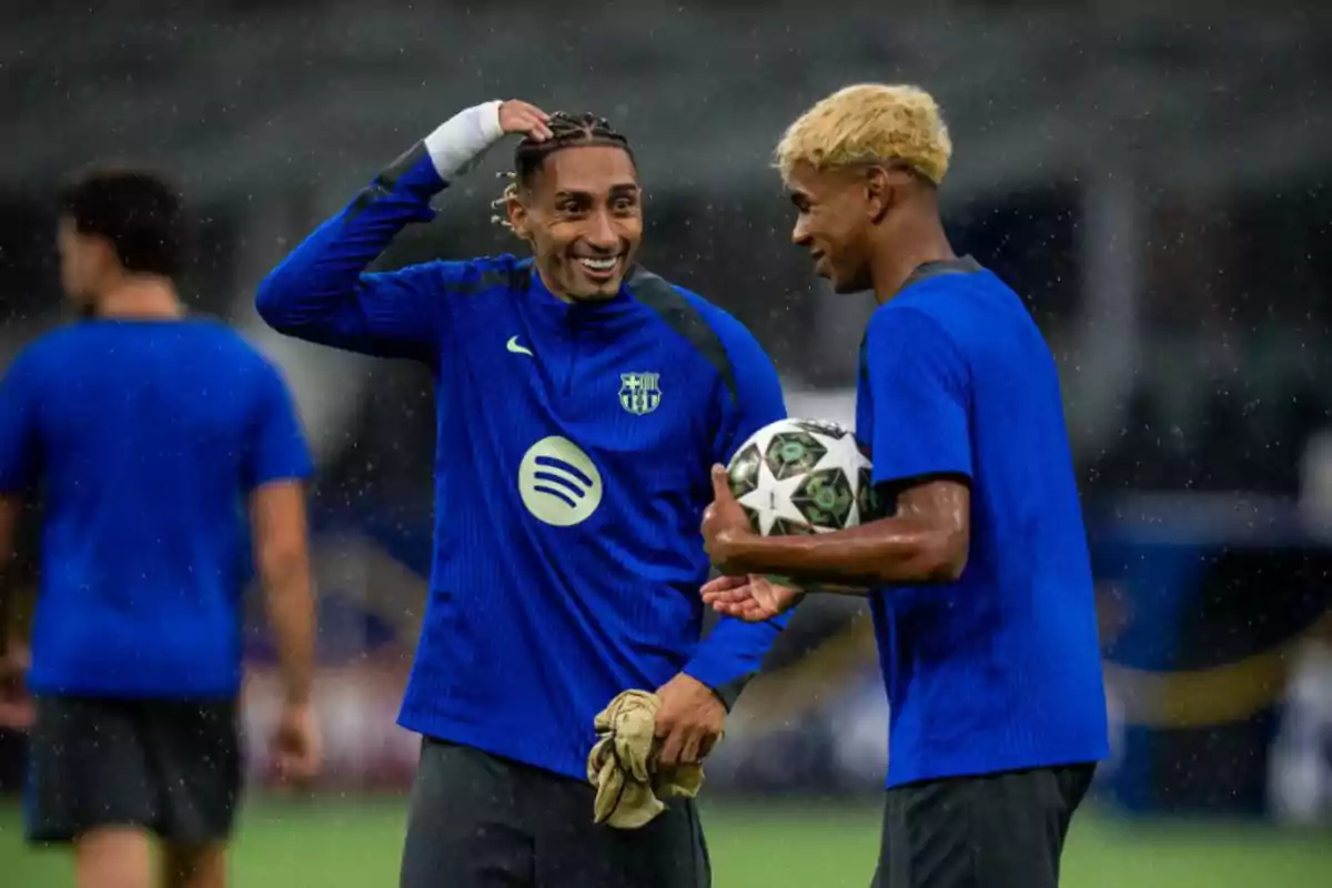 Two players in blue jerseys are talking and smiling on the field, one is holding a ball. Two players in blue jerseys are talking and smiling on the field, one is holding a ball.