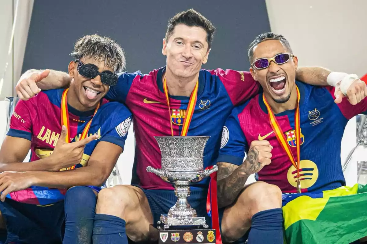 Trois footballeurs médaillés célèbrent avec un trophée, portant des t-shirts du FC Barcelone.