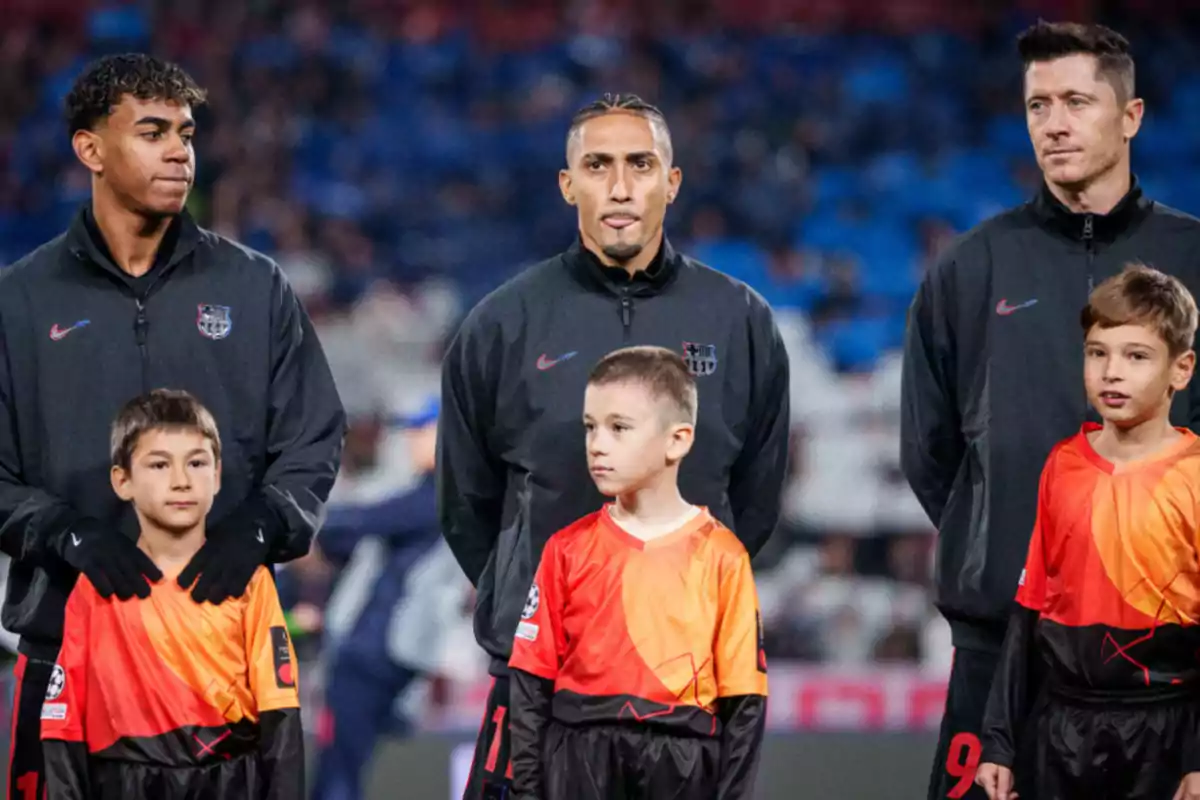 Jugadores de fútbol con chaquetas negras junto a niños con camisetas naranjas en un estadio.