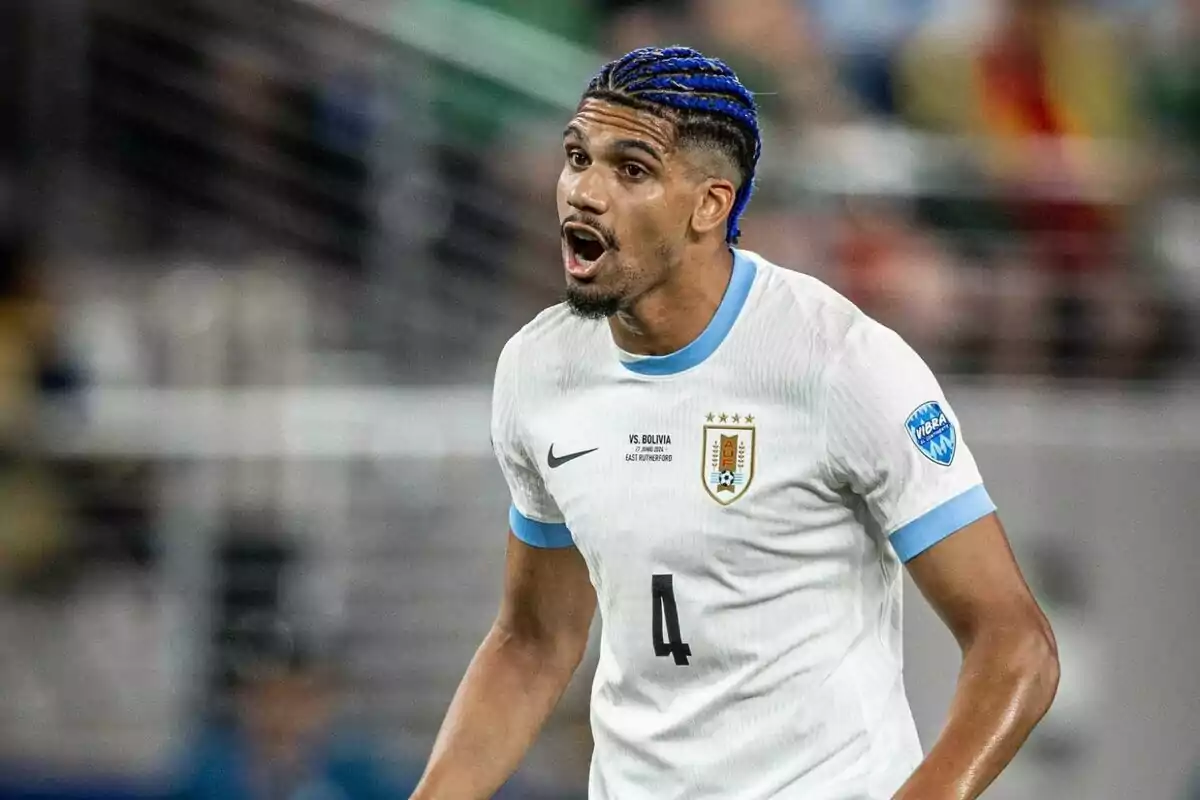 Jugador de fútbol con camiseta blanca de la selección de Uruguay y el número 4, con trenzas azules, en un estadio.