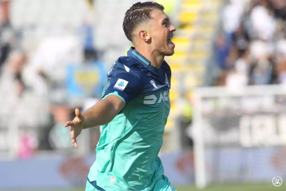 Samardzic celebrando un gol con los brazos abiertos y la lengua fuera, vestido con una camiseta azul marino y celeste del Udinese Calcio