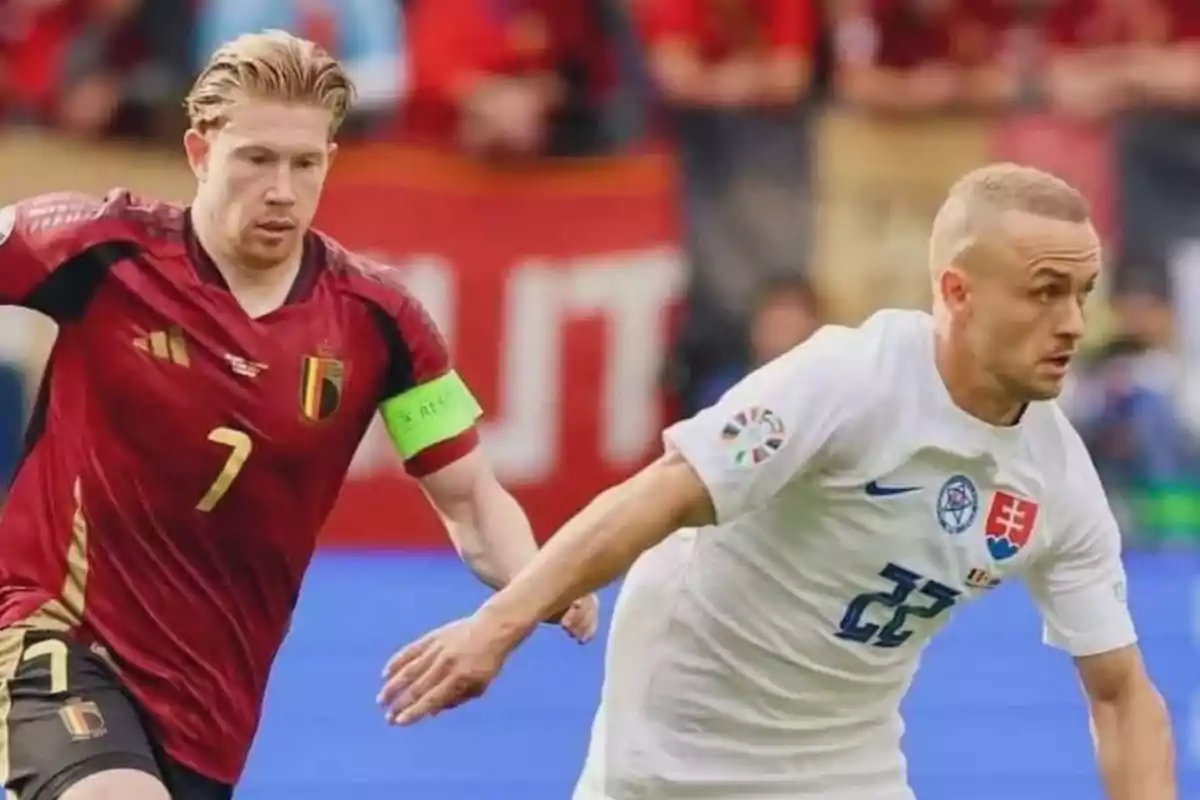 Dos jugadores de fútbol compiten por el balón durante un partido, uno de ellos lleva un uniforme rojo con el número 7 y un brazalete de capitán verde, mientras que el otro viste un uniforme blanco con el número 22.