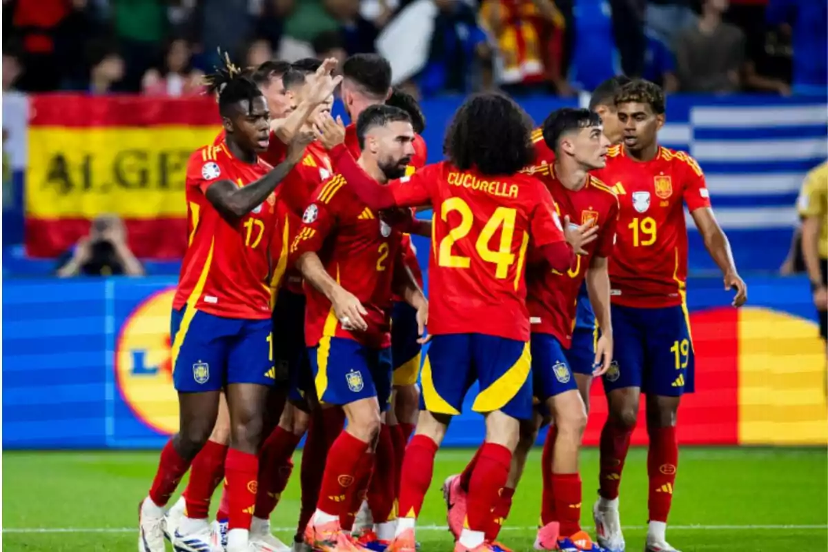 Jugadores de fútbol con uniforme rojo y amarillo celebran en el campo con una bandera de España de fondo.