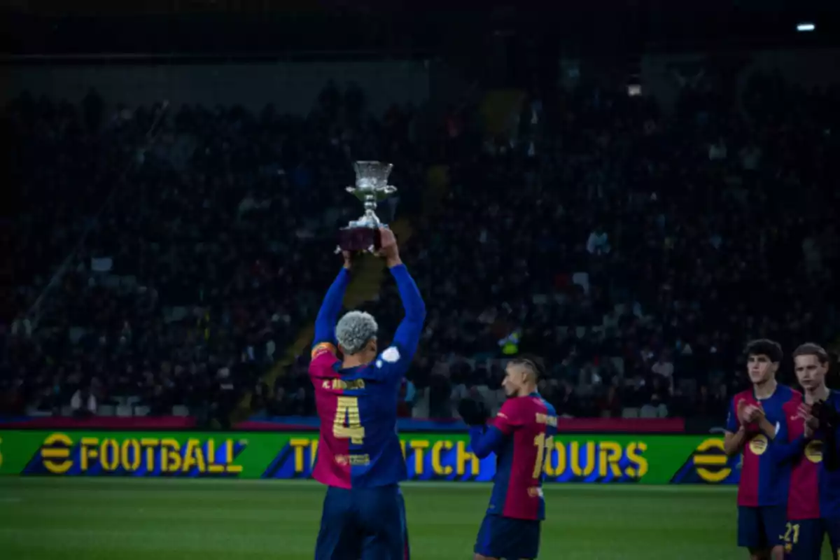 A soccer player lifts a trophy on the field as his teammates applaud and the crowd watches from the stands. A soccer player lifts a trophy on the field as his teammates applaud and the crowd watches from the stands.