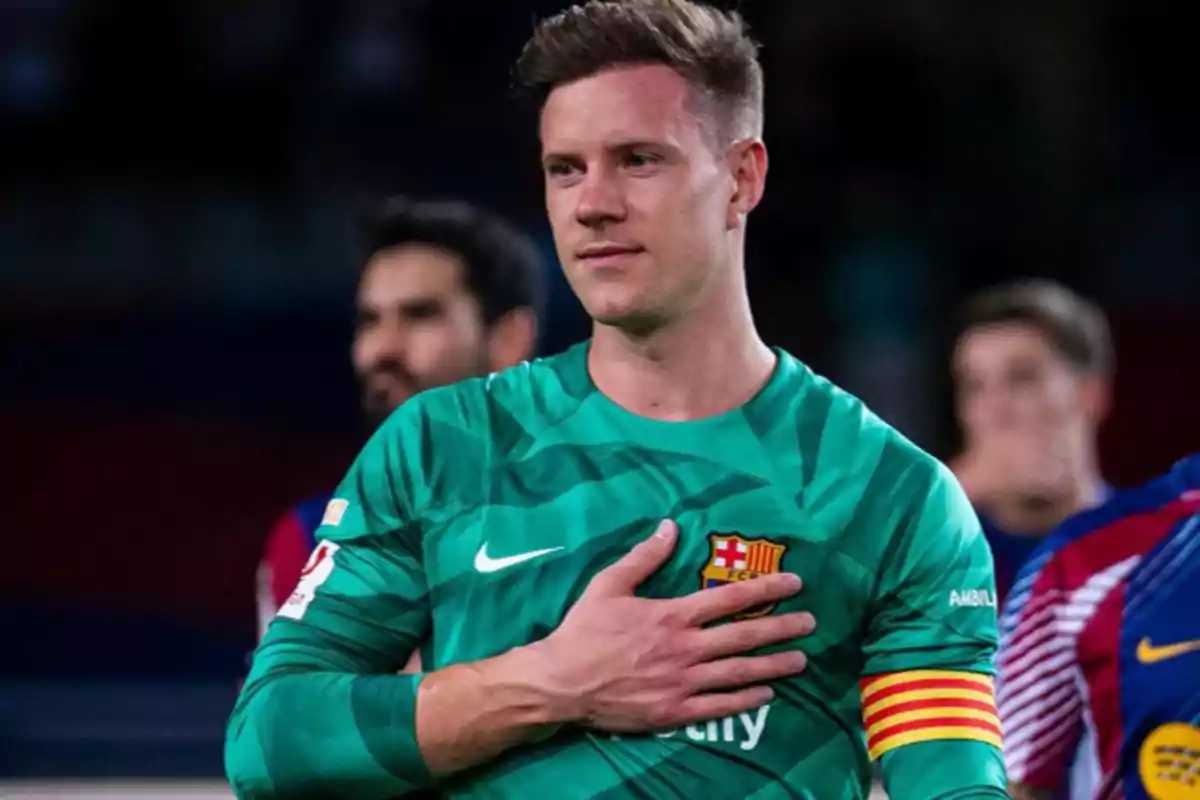 Un jugador de fútbol con uniforme verde y brazalete de capitán se lleva la mano al pecho.