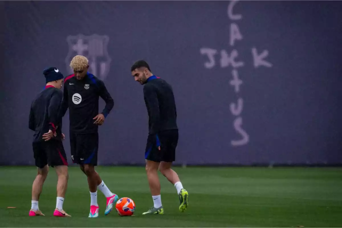Three players train on a grass field with a ball, wearing dark sportswear and brightly colored sneakers, while in the background a wall with the words Three players train on a grass field with a ball, wearing dark sportswear and brightly colored sneakers, while in the background a wall with the words