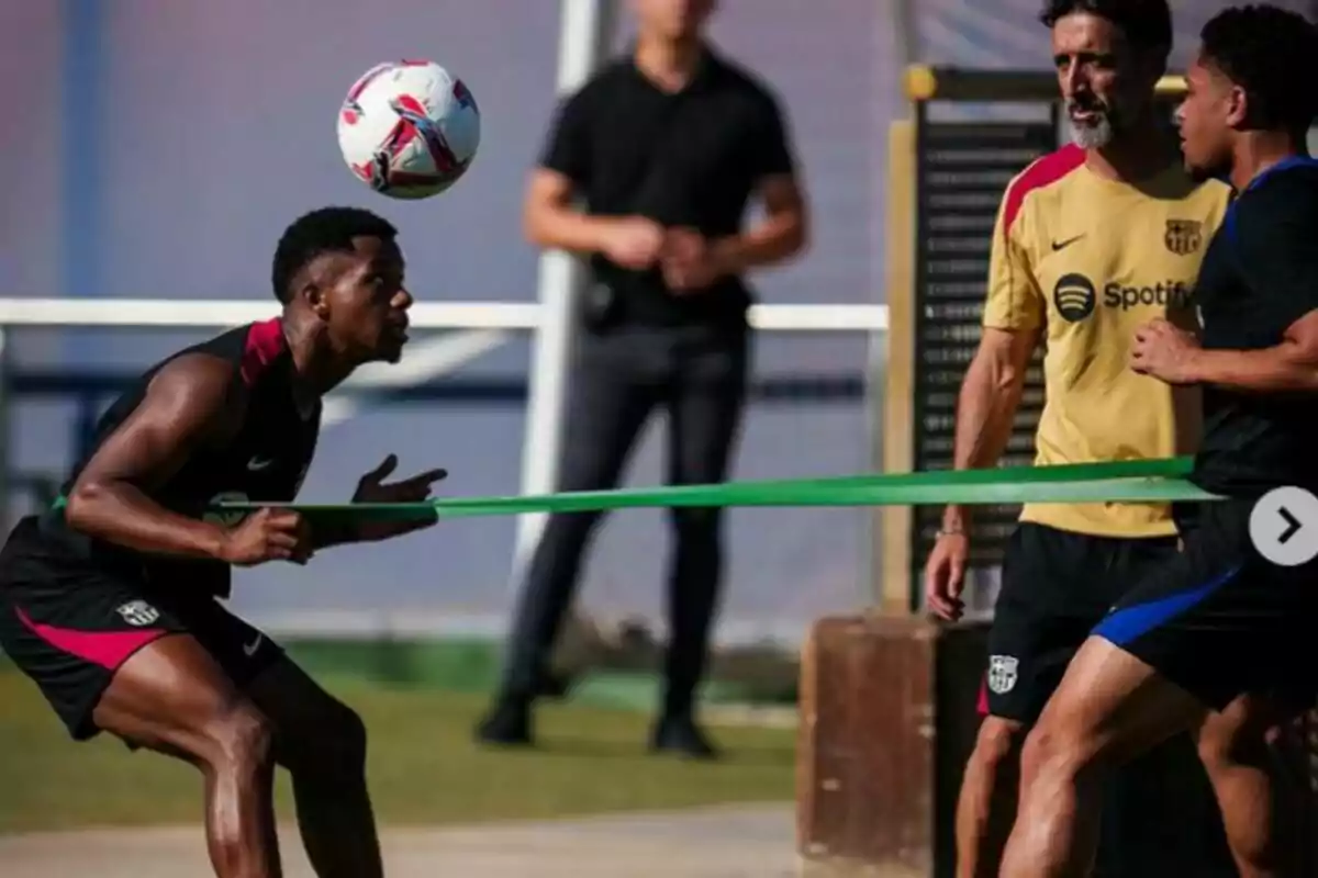 Jugadores de fútbol entrenando con una banda elástica mientras uno de ellos cabecea un balón, bajo la supervisión de un entrenador.