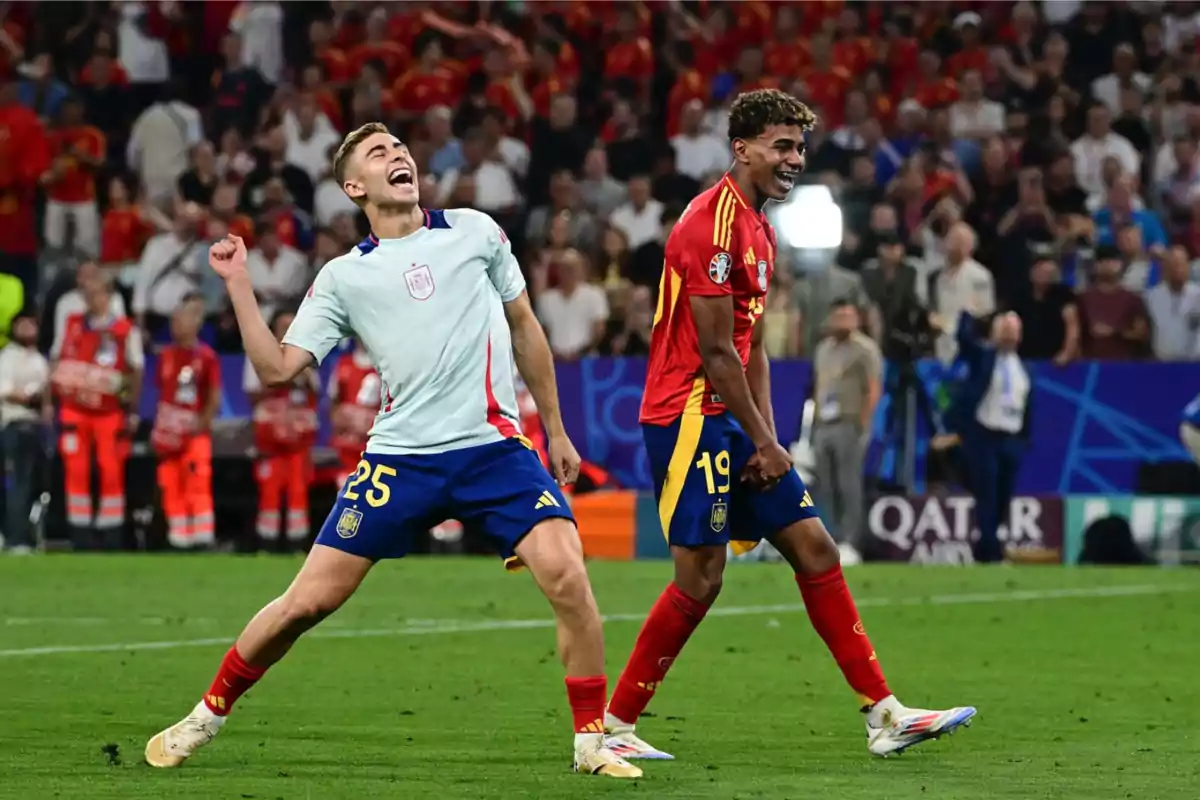 Dos jugadores de fútbol celebran en el campo mientras el público aplaude en el fondo.